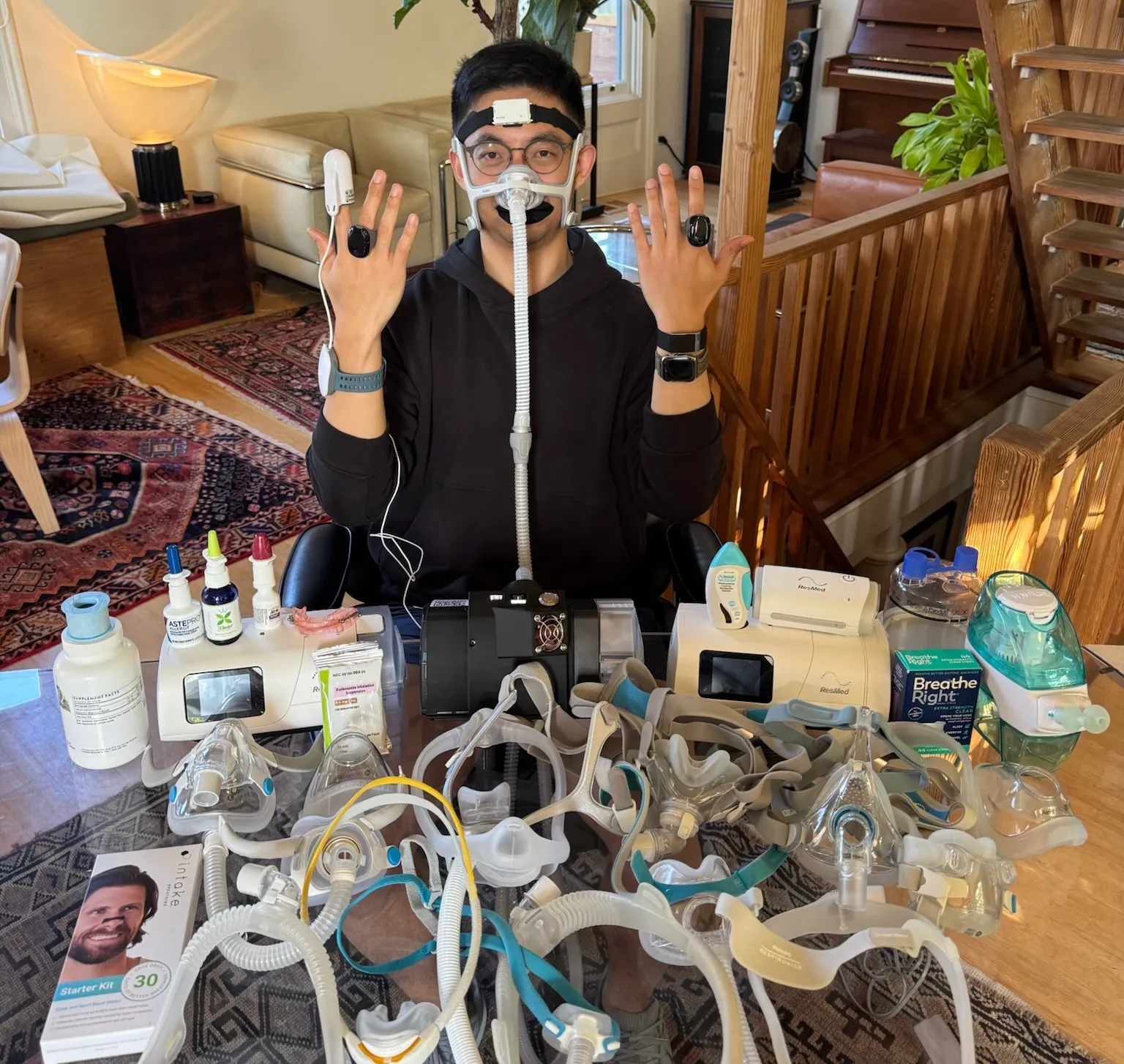 A Rhythm cofounder wearing a CPAP mask and finger sensors, seated behind a table covered with CPAP machines, masks, nasal sprays, and other sleep apnea treatments